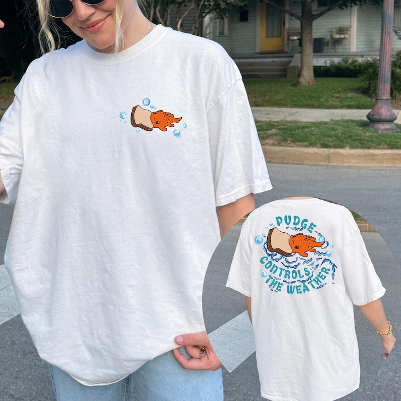 May include: White t-shirt with a cartoon design of an orange creature emerging from a brown object, surrounded by blue bubbles. The back of the shirt features the text "Pudge Controls The Weather" in blue.