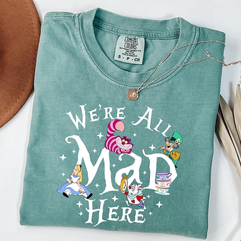 May include: A teal Comfort Colors t-shirt featuring the phrase "We're All Mad Here" with illustrations of characters from Alice in Wonderland. A silver necklace with a pendant is draped across the shirt.
