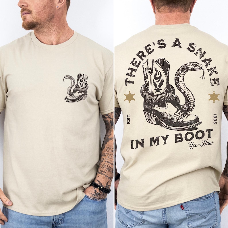 Pode incluir: Uma t-shirt bege de manga curta com um pequeno gr&aacute;fico preto de uma cobra enrolada numa bota de cowboy no peito esquerdo. As costas exibem uma vers&atilde;o grande do mesmo gr&aacute;fico com o texto THERE'S A SNAKE IN MY BOOT, EST. 1995, e Yee-Haw.