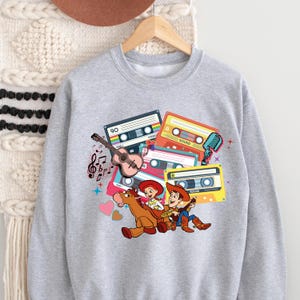 Puede incluir: Una sudadera gris claro con un gr&aacute;fico colorido de cintas de cassette, una guitarra, notas musicales y personajes de dibujos animados. El dise&ntilde;o incluye un micr&oacute;fono y el n&uacute;mero "90" en una de las cintas de cassette. Los personajes montan a caballo.
