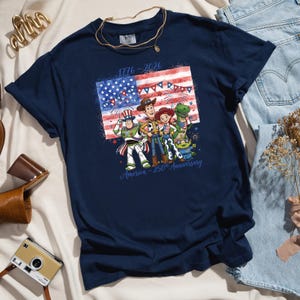 May include: Navy blue t-shirt featuring a graphic of the American flag and cartoon characters. The design includes the text "1776-2026" and "America - 250th Anniversary". The shirt is folded on a white surface with accessories.