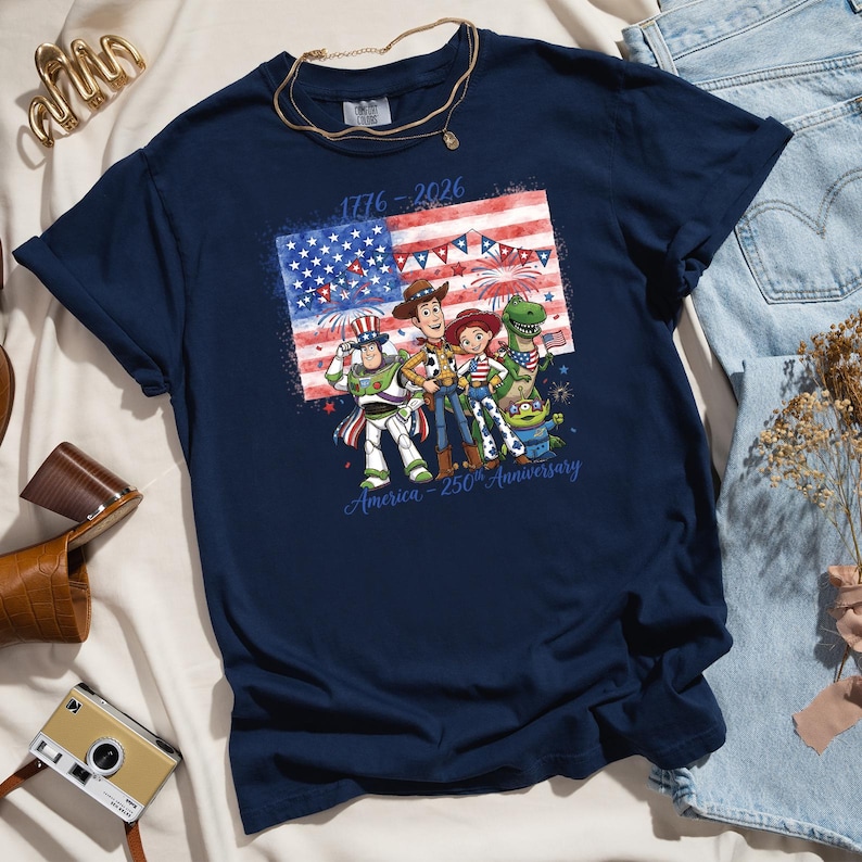 May include: Navy blue t-shirt featuring a graphic of the American flag and cartoon characters. The design includes the text "1776-2026" and "America - 250th Anniversary". The shirt is folded on a white surface with accessories.