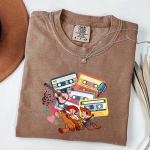 Puede incluir: Camiseta marr&oacute;n Comfort Colors con un gr&aacute;fico de cintas de cassette coloridas, una guitarra, notas musicales y personajes de dibujos animados. Tambi&eacute;n es visible un collar plateado con un colgante. La camiseta tiene una est&eacute;tica vintage y retro.