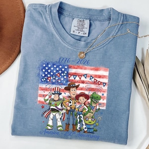 May include: A light blue t-shirt featuring a graphic of the American flag and cartoon characters. The shirt has the text "1776-2026" and "America, 250th Anniversary". A gold necklace is draped across the shirt.