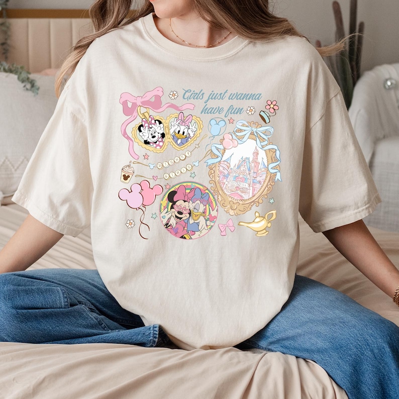 May include: Cream-colored t-shirt featuring a colorful graphic with Minnie Mouse, Daisy Duck, and a castle. The design includes pink bows, stars, and the text "Girls just wanna have fun."