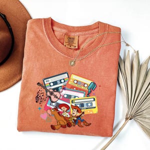 Puede incluir: Camiseta de color coral con un gr&aacute;fico de cintas de cassette, una guitarra, notas musicales y personajes de dibujos animados. La camiseta tiene cuello redondo y mangas cortas. Un collar dorado est&aacute; drapeado sobre la camiseta.