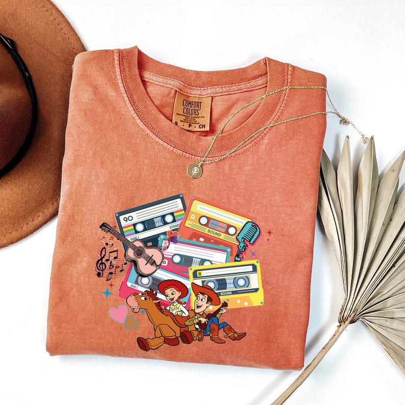 Puede incluir: Camiseta de color coral con un gr&aacute;fico de cintas de cassette, una guitarra, notas musicales y personajes de dibujos animados. La camiseta tiene cuello redondo y mangas cortas. Un collar dorado est&aacute; drapeado sobre la camiseta.