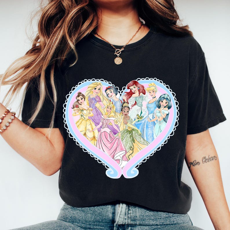 May include: Black t-shirt featuring a heart-shaped design with several cartoon princesses in colorful dresses. The heart has a light blue and pink border with a white scalloped edge. The princesses are in various poses.