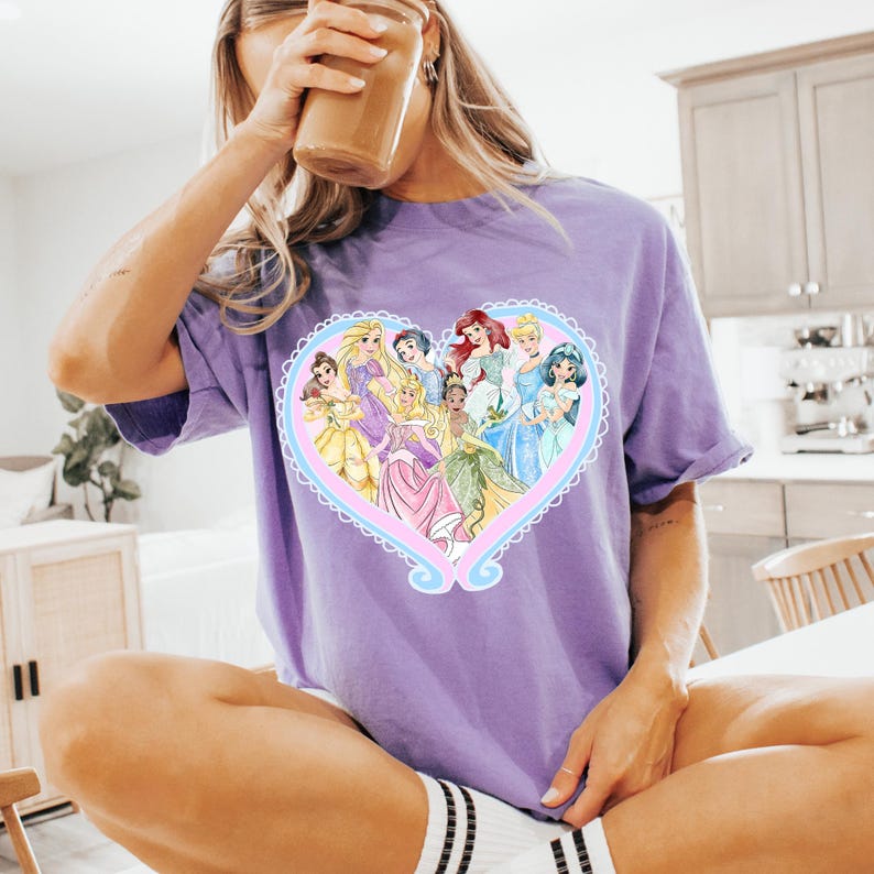 May include: A lilac-colored t-shirt featuring a heart-shaped design with several cartoon princesses. The heart has a light blue and pink border. The princesses are wearing colorful gowns. The shirt is made of a soft material.