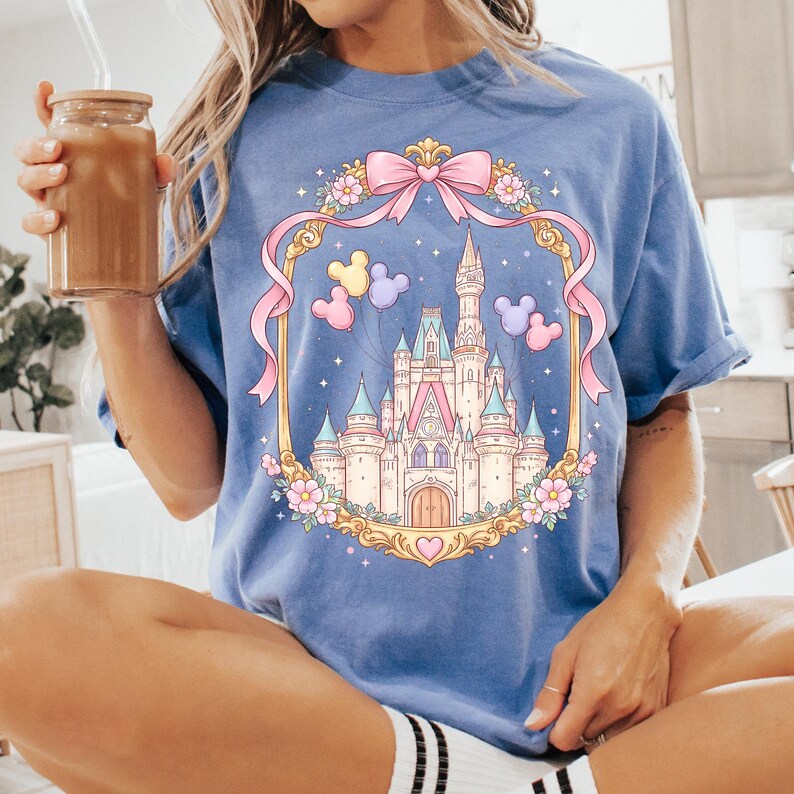 May include: A light blue t-shirt featuring a fairytale castle design within a gold frame with a pink bow and ribbons. The castle is surrounded by flowers and balloons. The shirt is made of soft material.