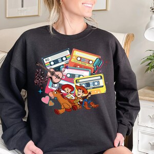 Puede incluir: Sudadera negra con un gr&aacute;fico colorido de cintas de cassette, una guitarra, notas musicales y personajes de dibujos animados. El dise&ntilde;o incluye el n&uacute;mero "90" y la palabra "SOUND" en las cintas. Una prenda divertida de tem&aacute;tica retro.