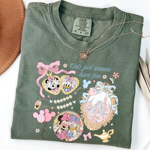 May include: Olive green t-shirt with a graphic featuring Minnie and Daisy Duck, heart-shaped sunglasses, and the phrase "Girls just wanna have fun." A gold necklace and a brown hat are also visible.