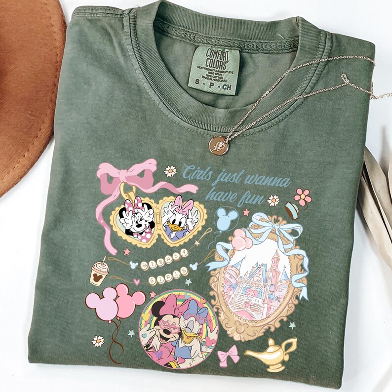 May include: Olive green t-shirt with a graphic featuring Minnie and Daisy Duck, heart-shaped sunglasses, and the phrase "Girls just wanna have fun." A gold necklace and a brown hat are also visible.