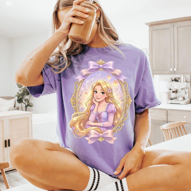 Op de afbeelding: Een lichtpaars T-shirt met een ingelijste illustratie van een tekenfilmprinses met lang blond haar en een paarse jurk. De lijst heeft een gouden rand en is versierd met strikken. De prinses glimlacht. Het shirt is een casual stijl.