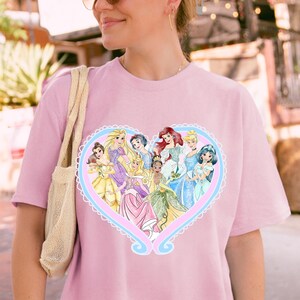 May include: A light pink t-shirt featuring a heart-shaped design with illustrations of various princesses. The heart is outlined in a light blue and white lace pattern. The princesses are depicted in their signature gowns.