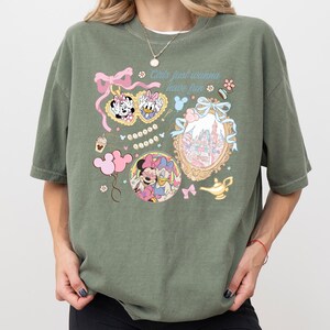 May include: An olive green t-shirt featuring cartoon graphics of Minnie Mouse and Daisy Duck, along with the text "Girls just wanna have fun." The design includes pink bows, a castle, and a magic lamp. The shirt has short sleeves and a relaxed fit.