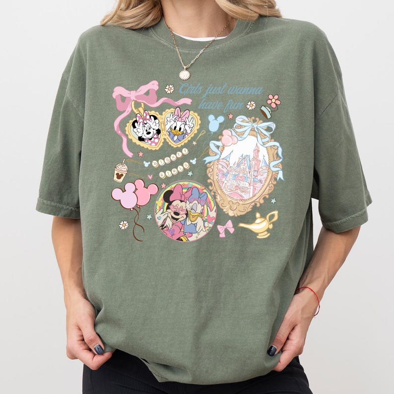 May include: An olive green t-shirt featuring cartoon graphics of Minnie Mouse and Daisy Duck, along with the text "Girls just wanna have fun." The design includes pink bows, a castle, and a magic lamp. The shirt has short sleeves and a relaxed fit.