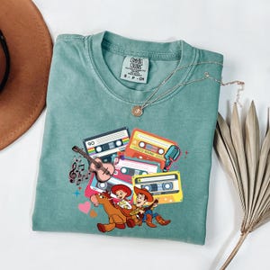 Puede incluir: Una camiseta Comfort Colors de color verde azulado con un gr&aacute;fico de cintas de cassette, una guitarra, notas musicales y personajes de dibujos animados. El dise&ntilde;o incluye el texto "SOUND" en las cintas de cassette. Un collar dorado est&aacute; sobre la camisa.