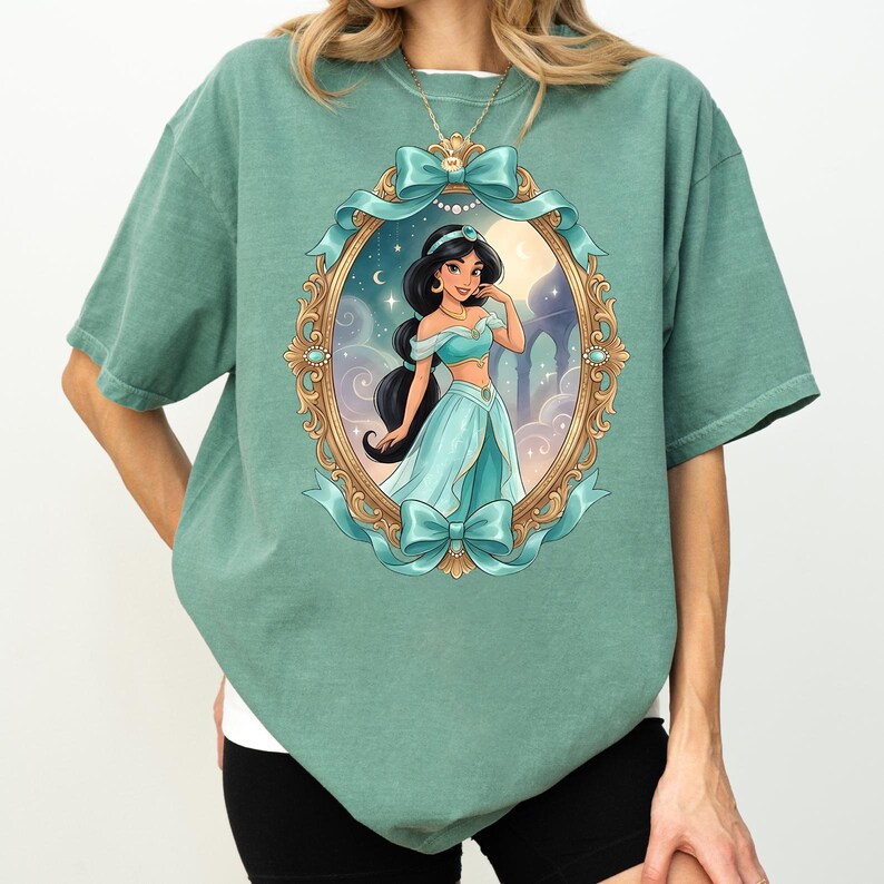 Op de afbeelding: Saliegroen t-shirt met een ingelijste illustratie van een prinses in een turquoise jurk. De gouden lijst heeft turquoise strikken en linten. De prinses heeft lang zwart haar en een nachthemel en paleis achtergrond.
