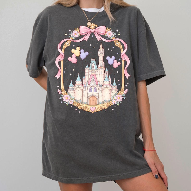 May include: Dark gray t-shirt featuring a colorful illustration of a fairytale castle within a decorative frame. The frame includes a pink bow, flowers, and Mickey Mouse balloons. The castle is pink, blue, and gold.