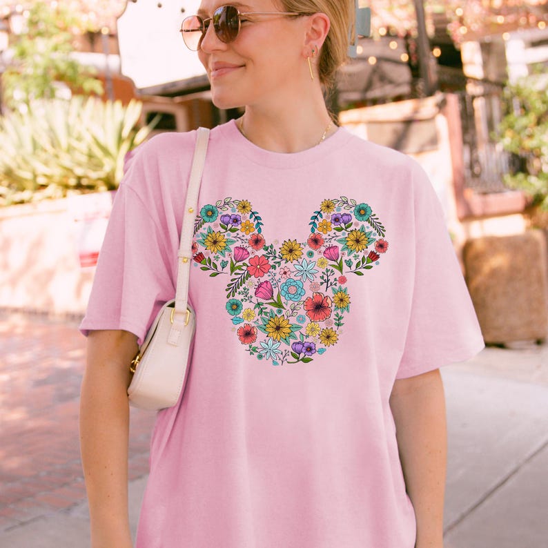 K&ouml;nnte beinhalten: Ein hellrosa T-Shirt mit einem floralen Mickey Mouse-Design. Das Design besteht aus verschiedenen bunten Blumen, darunter rosa, gelbe, blaue und violette. Das Shirt hat kurze &Auml;rmel und einen Rundhalsausschnitt.