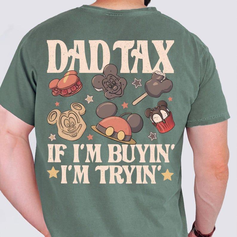 May include: Green t-shirt with the text "DAD TAX" above a graphic of Mickey Mouse-themed treats. Below the treats, the text reads "IF I'M BUYIN' I'M TRYIN'" with star accents. The shirt is a solid color.