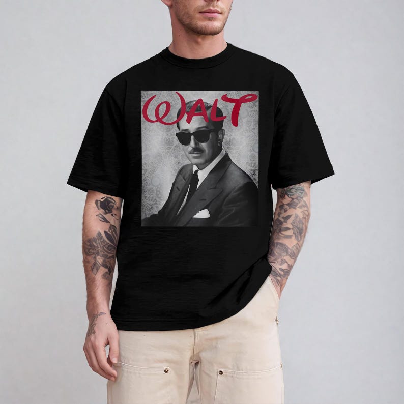 May include: Black t-shirt featuring a black and white portrait of a man wearing sunglasses and a suit. The word "WALT" is written in red cursive above the portrait. The shirt is made of a soft material.