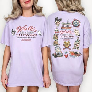 May include: A light purple t-shirt featuring the text "Walt's Ink & Paint Tattoo Shop" in a vintage style. The shirt has various tattoo-style graphics on the front and back, including Mickey Mouse imagery and other designs. The text "The Most Magical Tattoos on Earth!" is also present.