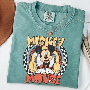 May include: A teal t-shirt with a Mickey Mouse graphic. The design features the words "Mickey Mouse" in yellow and red, with Mickey in the center. A gold necklace with a pendant is draped across the shirt.