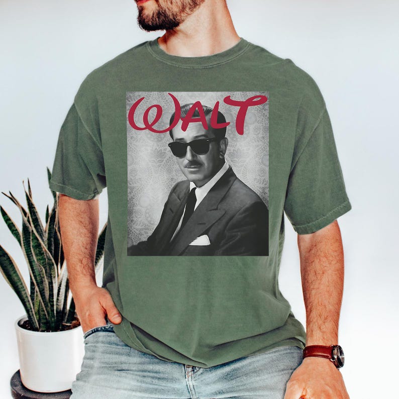 May include: Olive green t-shirt featuring a black and white image of a man wearing sunglasses and a suit. The word "WALT" is written in red cursive above the image. The shirt is a casual style.