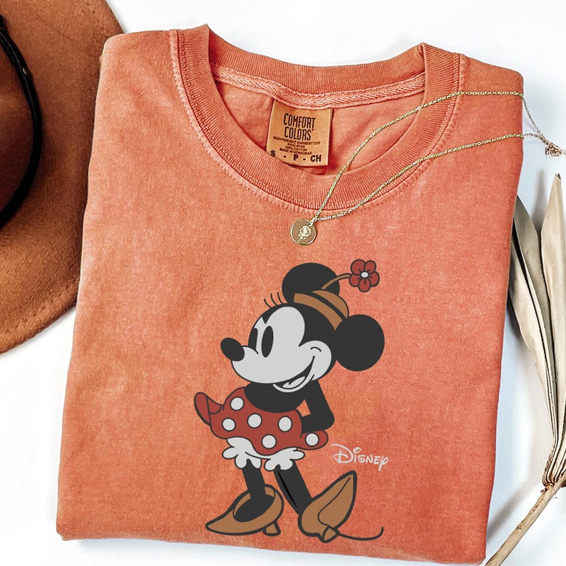 Może przedstawiać: Pomarańczowa koszulka Comfort Colors z grafiką Myszki Minnie. Postać z kresk&oacute;wki ma na sobie czerwoną sukienkę w kropki, brązowe buty i kapelusz z kwiatkiem. Złoty naszyjnik jest zarzucony na koszulkę.