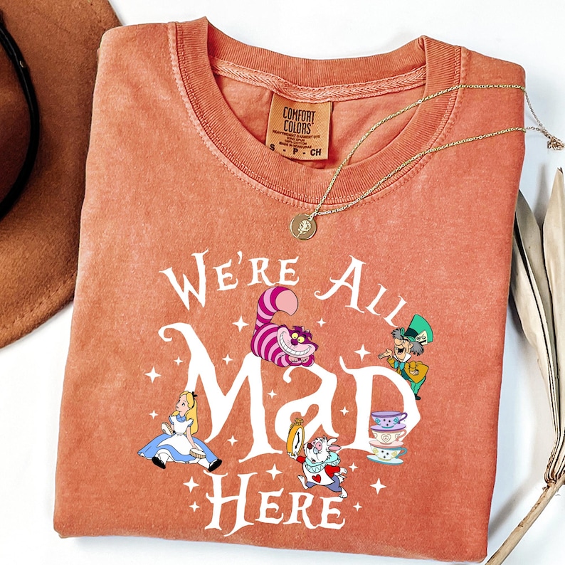 May include: Coral-colored Comfort Colors t-shirt featuring the phrase "We're All Mad Here" with Alice in Wonderland characters. A gold necklace with a pendant is draped across the shirt. A brown hat is visible in the upper left corner.