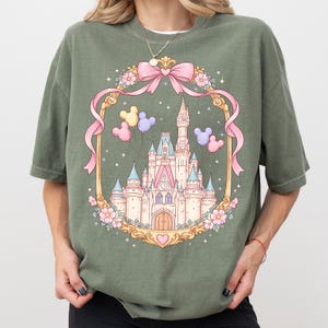 May include: Olive green t-shirt featuring a fairytale castle design within a gold frame adorned with pink ribbons, flowers, and Mickey Mouse balloons. The castle has pink and blue turrets and a heart at the base.
