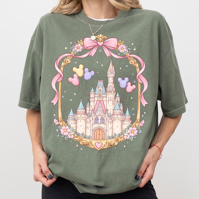 May include: Olive green t-shirt featuring a fairytale castle design within a gold frame adorned with pink ribbons, flowers, and Mickey Mouse balloons. The castle has pink and blue turrets and a heart at the base.