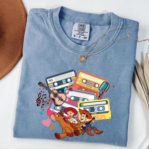 Puede incluir: Una camiseta azul claro con un gr&aacute;fico colorido de cintas de cassette, una guitarra, notas musicales y personajes de dibujos animados. La camiseta tiene cuello redondo y mangas cortas. Tambi&eacute;n se ve un collar dorado con un colgante.