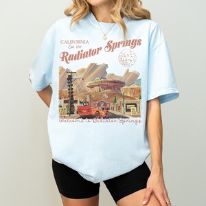 May include: Light blue t-shirt featuring a graphic of Radiator Springs, California, with the text "Radiator Springs" and "Welcome to Radiator Springs." The design includes a cartoon car and desert landscape.