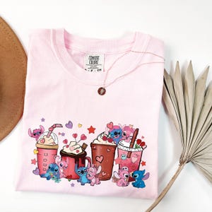 Puede incluir: Camiseta rosa p&aacute;lido Comfort Colors con un gr&aacute;fico de personajes de dibujos animados, bebidas de caf&eacute; y corazones. Un delicado collar con un colgante descansa sobre la camiseta. Un sombrero de tonos neutros y una fronda de palma seca est&aacute;n en el fondo.