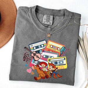 Puede incluir: Una camiseta gris Comfort Colors con un gr&aacute;fico colorido de cintas de cassette, una guitarra, notas musicales y personajes de dibujos animados. La camiseta tiene una peque&ntilde;a etiqueta en el cuello y un collar plateado.