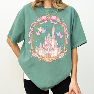 May include: Sage green t-shirt featuring a fairytale castle design within a gold frame. The design includes a pink bow, Mickey Mouse balloons, and floral accents. The castle has pink and blue turrets.