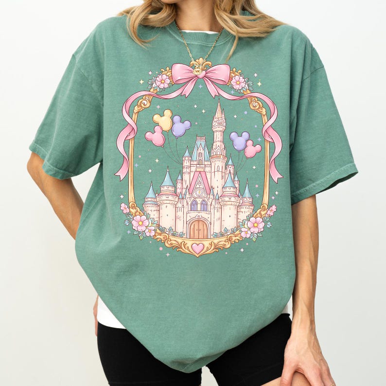 May include: Sage green t-shirt featuring a fairytale castle design within a gold frame. The design includes a pink bow, Mickey Mouse balloons, and floral accents. The castle has pink and blue turrets.