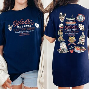 May include: Navy blue t-shirt featuring the text "Walt's Ink & Paint Tattoo Shop" with various tattoo-style graphics. The front of the shirt displays the shop's name and logo, while the back showcases multiple tattoo designs, including a skull, boat, and Mickey Mouse hand.