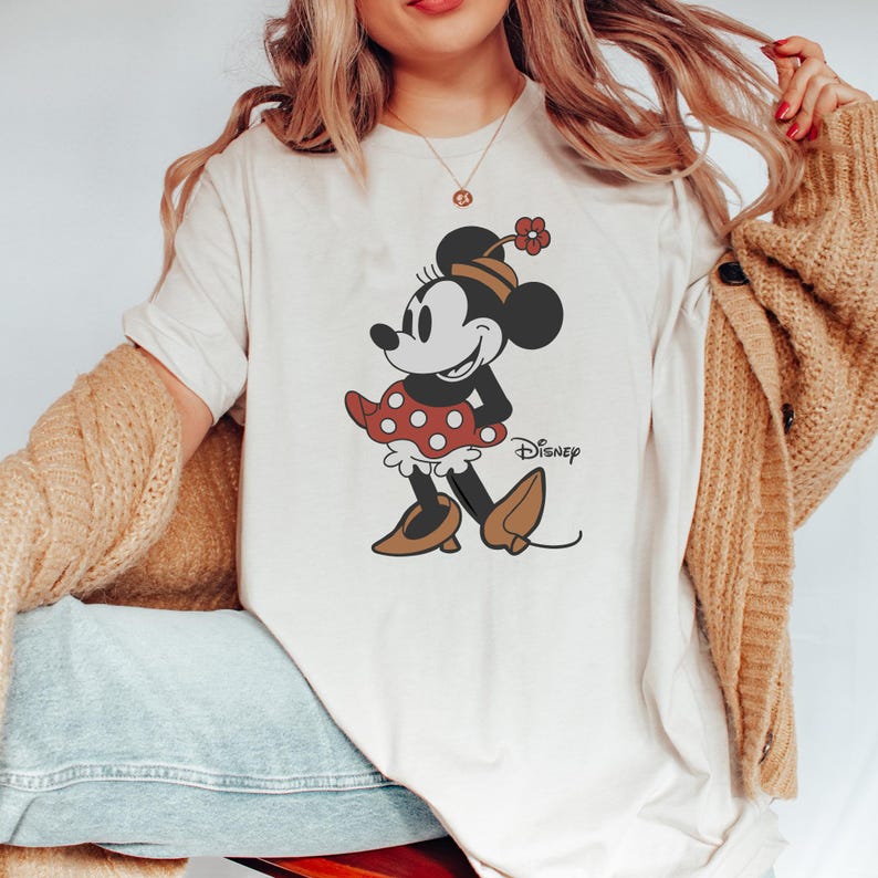 Może przedstawiać: Kremowy t-shirt z grafiką Myszki Minnie. Myszka Minnie jest przedstawiona w swojej kultowej czerwonej sukience w kropki, brązowych butach i kwiatkiem na kapeluszu. Poniżej znajduje się logo Disney.