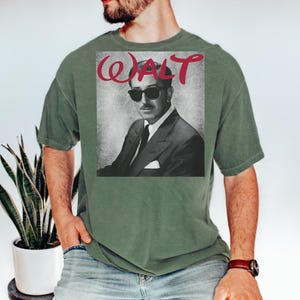 May include: Olive green t-shirt featuring a black and white image of a man in a suit and sunglasses, with the name "WALT" in red script above the image. The shirt has short sleeves and a crew neck.