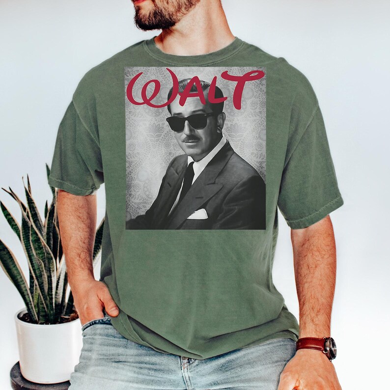 May include: Olive green t-shirt featuring a black and white image of a man in a suit and sunglasses, with the name "WALT" in red script above the image. The shirt has short sleeves and a crew neck.