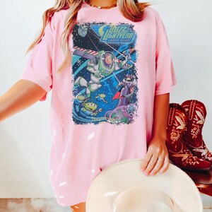 K&ouml;nnte beinhalten: Rosa T-Shirt mit einem Aufdruck von Buzz Lightyear aus dem Film Toy Story. Der Aufdruck zeigt den Schriftzug "Buzz Lightyear Laser Blast" und Abbildungen der Charaktere. Dazu rote Cowboy-Stiefel.