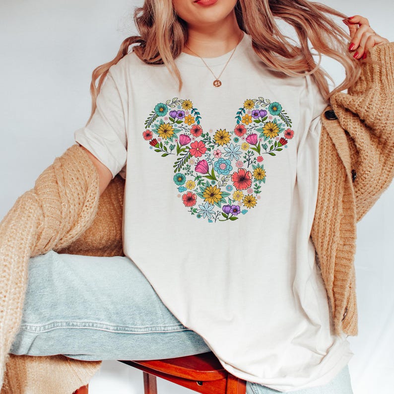 K&ouml;nnte beinhalten: Cremefarbenes T-Shirt mit einem floralen Mickey-Mouse-Design. Das Design besteht aus verschiedenen bunten Blumen, darunter rosa, gelbe, blaue und lila, die in Form von Mickey Mouses Kopf und Ohren angeordnet sind. Das Shirt ist ein l&auml;ssiger, bequemer Stil.