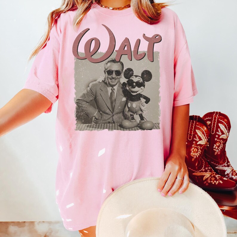 May include: Pink t-shirt featuring a vintage-style graphic of Walt Disney and Mickey Mouse. The word "Walt" is printed above the image in a cursive font. The shirt is paired with red cowboy boots.