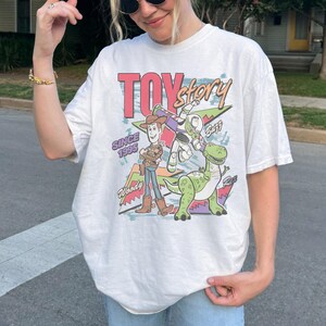 May include: White t-shirt featuring a colorful graphic of characters from the movie "Toy Story." The design includes Woody, Buzz Lightyear, and Rex, with the text "TOY STORY" and "SINCE 1995." The shirt has a vintage, retro style.