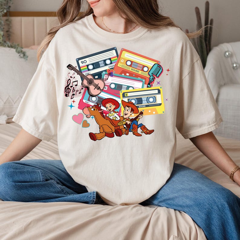 Puede incluir: Camiseta color crema con un gr&aacute;fico colorido de cintas de cassette, una guitarra, notas musicales y personajes de dibujos animados. El dise&ntilde;o incluye un vaquero y una vaquera montados a caballo. La camiseta tiene mangas cortas y un corte holgado.