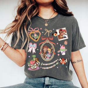 May include: Dark grey t-shirt with colourful cartoon graphics of Toy Story characters. The design includes images of Woody, Buzz Lightyear, and other characters, along with bows and stars.